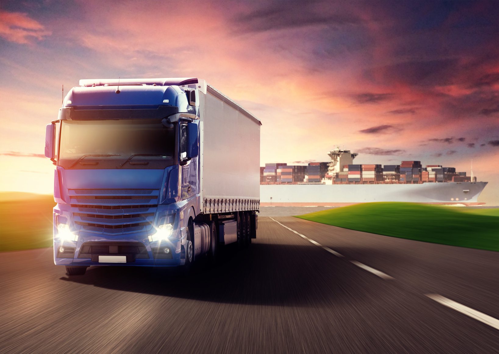 A large blue cargo truck drives on an open highway under a dusk sky with vibrant colors. In the background, a container ship loaded with shipping containers sails on a distant body of water. The scene depicts transportation and logistics.